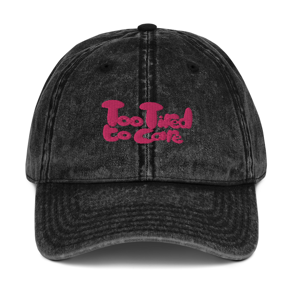 The front of a black vintage cotton twill cap with a pink slogan on the from that reads "Too Tired to Care".