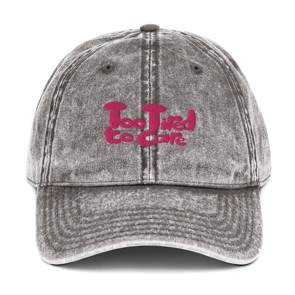 The front of a vintage charcoal grey cotton twill cap with a pink slogan on the front that reads "Too Tired to Care".