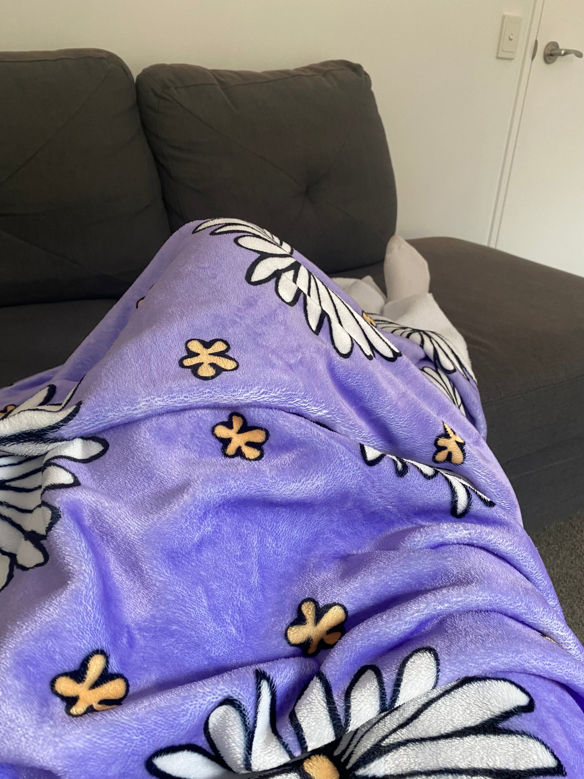 A photo of a purple blanket patterned with hand-drawn white daisies with yellow centres and miniature hand-drawn yellow flowers. The photo shows a person's legs covered in the blanket while they sit on a grey sofa, with their feet poking out from the bottom of the blanket.