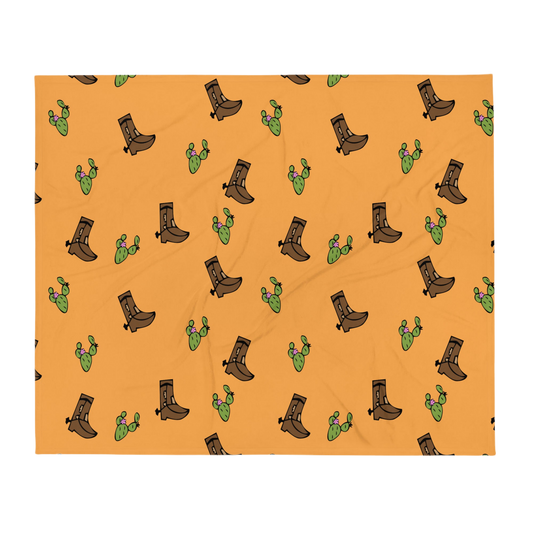 A photo of a vibrant orange blanket patterned with hand-drawn cowboy boots in shades of brown and hand-drawn green cacti with pink flowers.