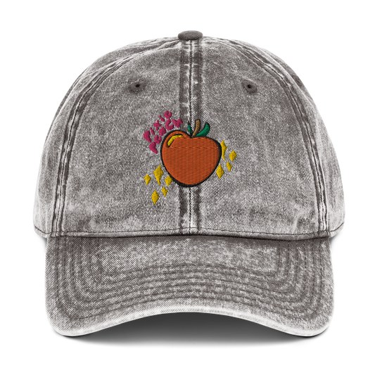 A photo of the front of a charcoal grey cotton twill cap, showcasing the Pixie Peach logo (pink text, orange peach with green leaves and a brown stalk, yellow sparkles and yellow reflective gleam on the peach). 