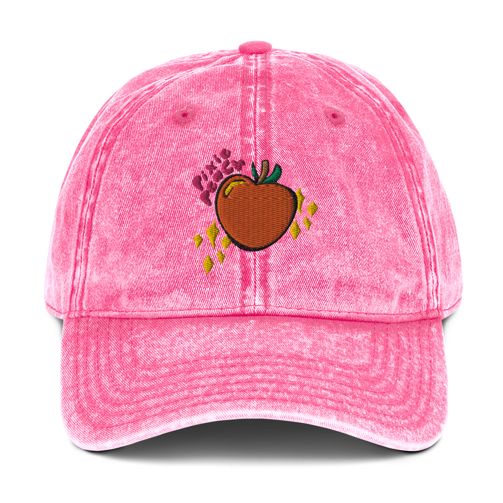 A photo of the front of a pink cotton twill cap, showcasing the Pixie Peach logo (pink text, orange peach with green leaves and a brown stalk, yellow sparkles and yellow reflective gleam on the peach). 